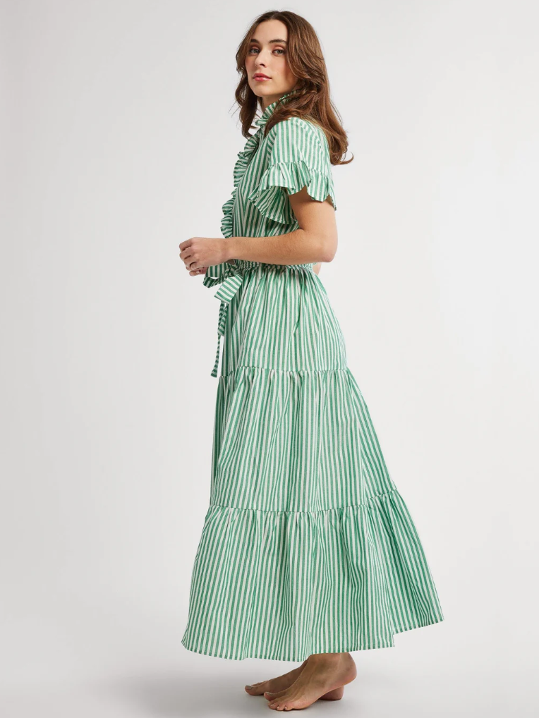 A woman wearing a green and white striped maxi dress with short sleeves and a tie at the waist, walking gracefully to the left, against a light gray background in Scottsdale, Arizona. The dress is the Victoria Dress by Mille.