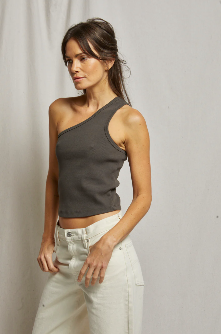 A woman poses in front of a plain background at her bungalow, wearing a Perfectwhitetee Call Me One Shoulder Structured Rib Tank and white jeans, looking away from the camera with a thoughtful expression.
