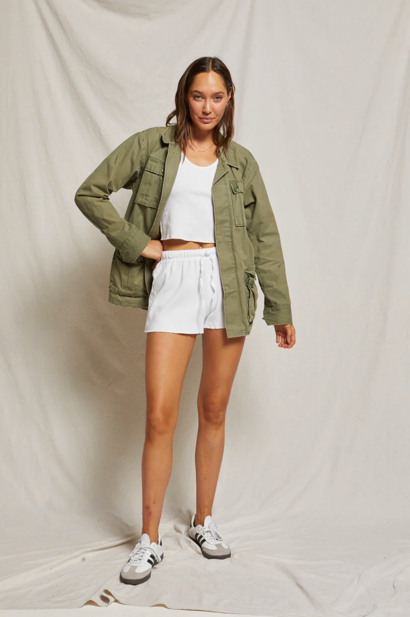 A young woman stands confidently against a beige backdrop in a bungalow, wearing a white tank top, olive green jacket, and Graham Waffle Shorts from Perfectwhitetee, complemented by gray sneakers.