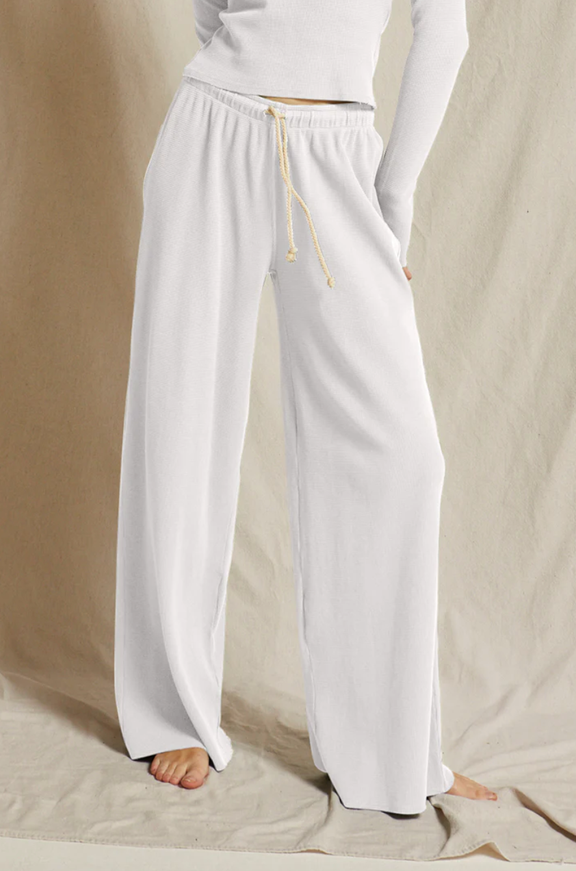 A person stands wearing Perfectwhitetee's Rivers Waffle Wide-Leg pants and a long-sleeve white top, with a bungalow-style beige backdrop. The focus is on the casual, relaxed fit of the pants.