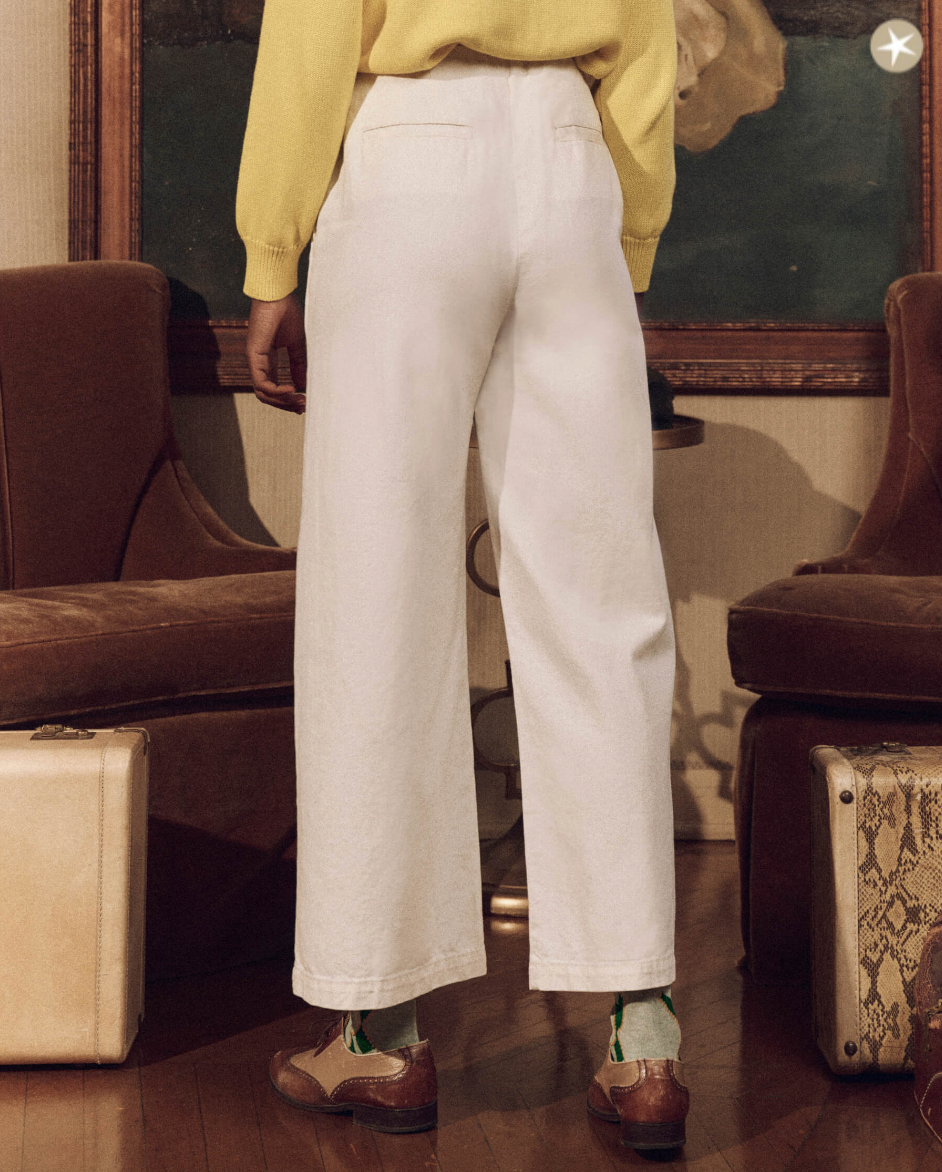 A person in a yellow sweater and The Great Inc.'s Sculpted Trouser stands in a vintage bungalow with wood furnishings, facing away from the camera. Focus is on the clothing style and setting.
