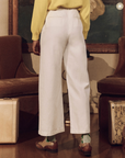 A person in a yellow sweater and The Great Inc.'s Sculpted Trouser stands in a vintage bungalow with wood furnishings, facing away from the camera. Focus is on the clothing style and setting.