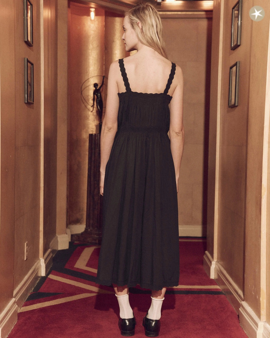 A woman in a black Victorian dress (The Cachet Dress from The Great Inc.) and white socks with black shoes walks away down a warmly lit corridor with red and green carpet. Her blonde hair is pulled up loosely.