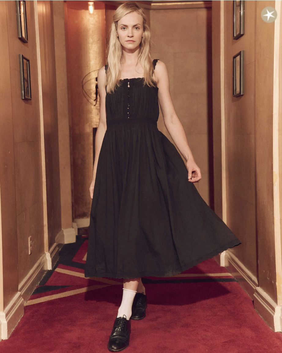 A woman with blonde hair stands in a hallway wearing a black, mid-length scalloped lace Cachet dress from The Great Inc. and black shoes with white socks, looking directly at the camera with a neutral expression.