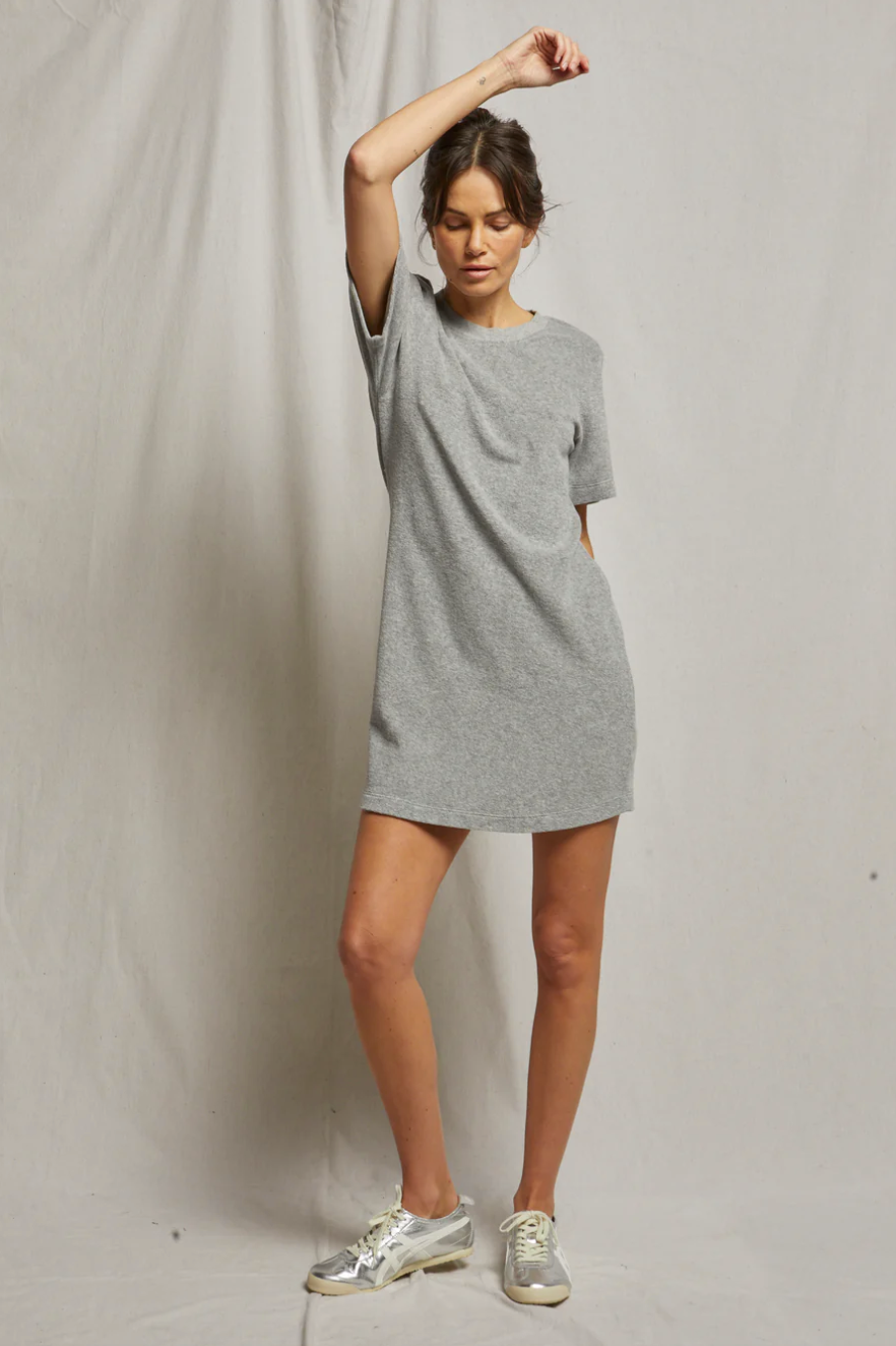A woman in a Perfectwhitetee EMELIA loop terry tee dress and silver sneakers poses with one arm raised, standing against a draped light background in a bungalow. Her pose is relaxed and elegant.