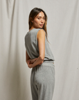 A woman with long brown hair stands sideways, looking over her shoulder at the camera. She wears the Perfectwhitetee ELLIE terry banded tank, paired with matching gray pants, against a neutral fabric backdrop.