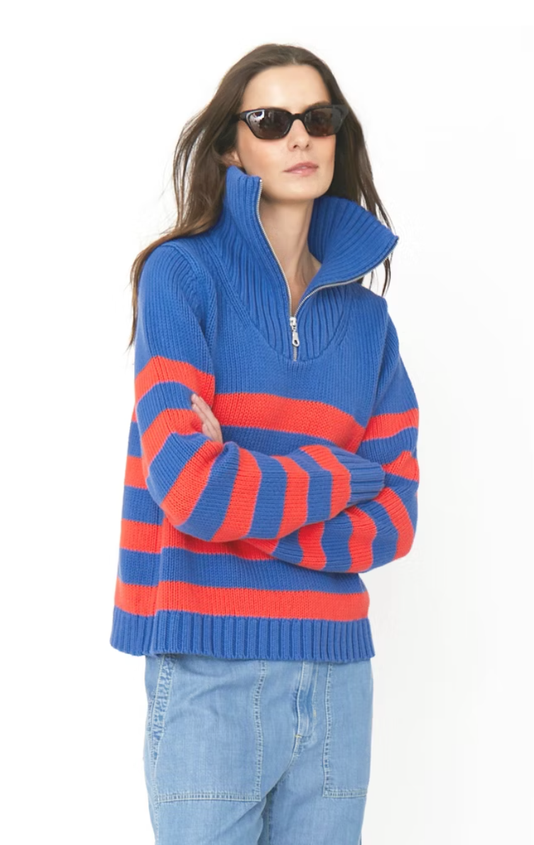 A woman in sunglasses stands with her arms crossed, wearing a blue and orange striped cotton quarter zip sweater (The Matey by Kule) and light blue jeans against a white background.