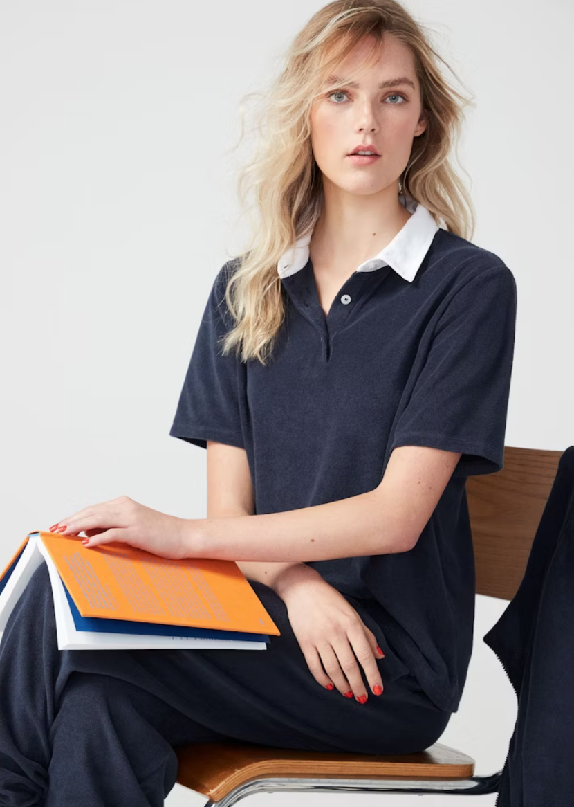 A woman with blond hair sits on a wooden stool, holding an orange notebook. She wears a machine washable, navy blue Kule terry polo shirt with a white collar and looks thoughtfully at the camera.
