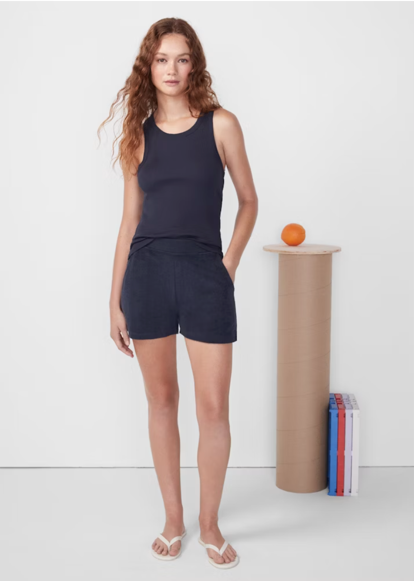 A woman with curly hair stands wearing a navy tank top and Kule's Terry Venus shorts, posing next to a pedestal with an orange on top and books beside it against a white backdrop.
