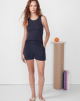 A woman with curly hair stands wearing a navy tank top and Kule's Terry Venus shorts, posing next to a pedestal with an orange on top and books beside it against a white backdrop.
