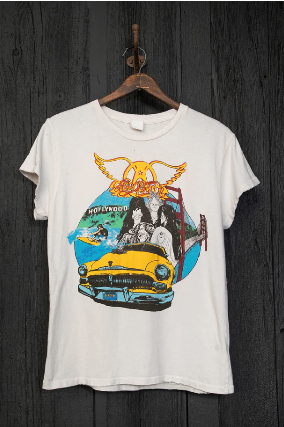 A Made Worn Aerosmith 1977 tour vintage white T-shirt with a colorful graphic design hanging on a wooden hanger against a dark wooden background. The design features a classic car and Hollywood-themed illustrations, including subtle elements inspired by Scottsdale Arizona.