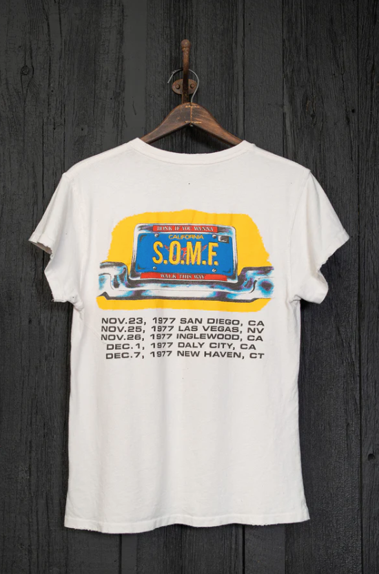 A vintage white Made Worn T-shirt hanging on a wooden hanger against a dark wood background, featuring a colorful print of a van and tour dates from 1977 in various US cities including Scottsdale, Arizona.