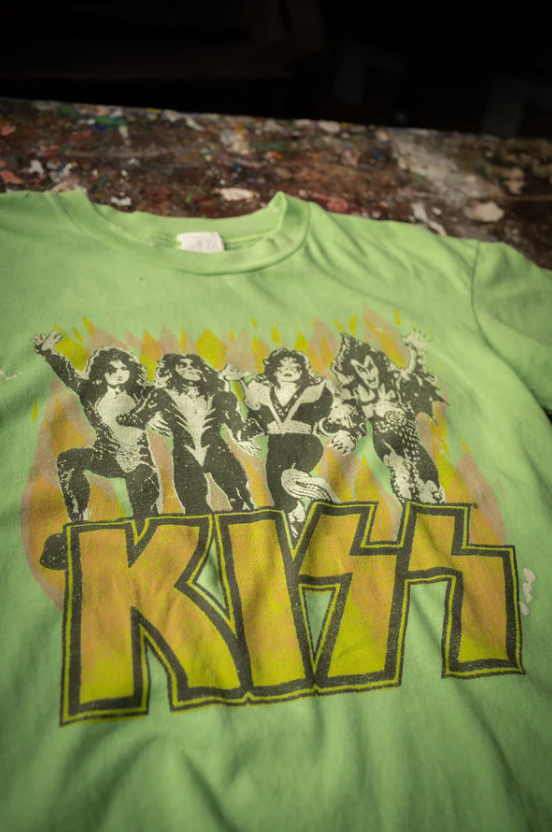 A vintage green Made Worn Kiss Crop Tee Electric featuring a graphic print of the band members in dramatic poses above the bold, yellow "KISS" logo. The shirt is displayed on a textured, Arizona-style surface.