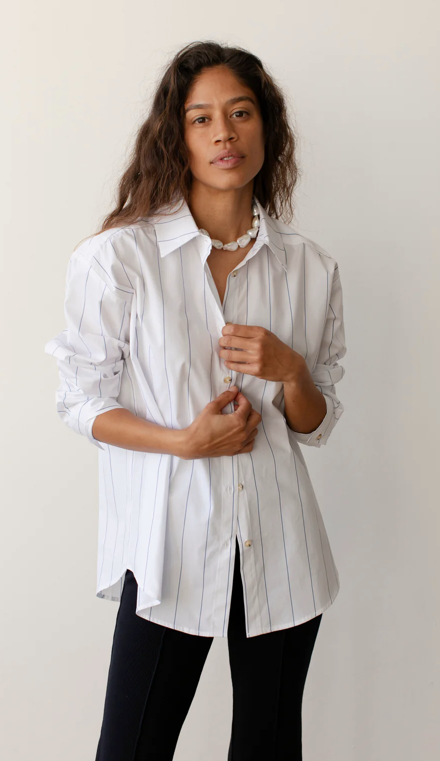 A woman stands against a plain background, wearing a white Donni Stripe Pop Shirt and black pants, featuring tortoise-like buttons. She is buttoning her cuffs. She has curly brown hair and a serene expression.