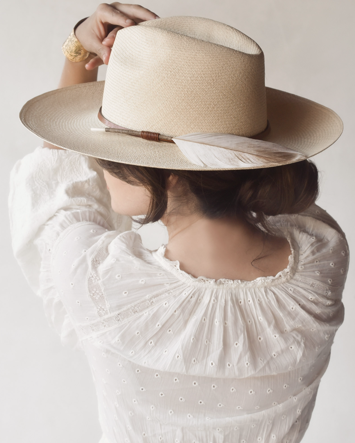 A woman in a white blouse with a ruffled neckline is seen from behind, holding a stylish Ninakuru oquilla straw hat over her head. Her brown hair is neatly tucked under the Matteo Extra Long Brim S/M hat.