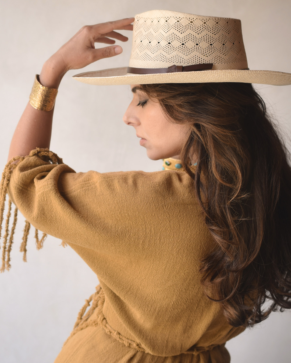 A woman in profile, wearing a Ninakuru Kaela Extra Long Brim M hat crafted from artisanal Toquilla straw and a textured mustard dress with fringe details, touches the rim of her hat. Her wavy hair cascades down her back.
