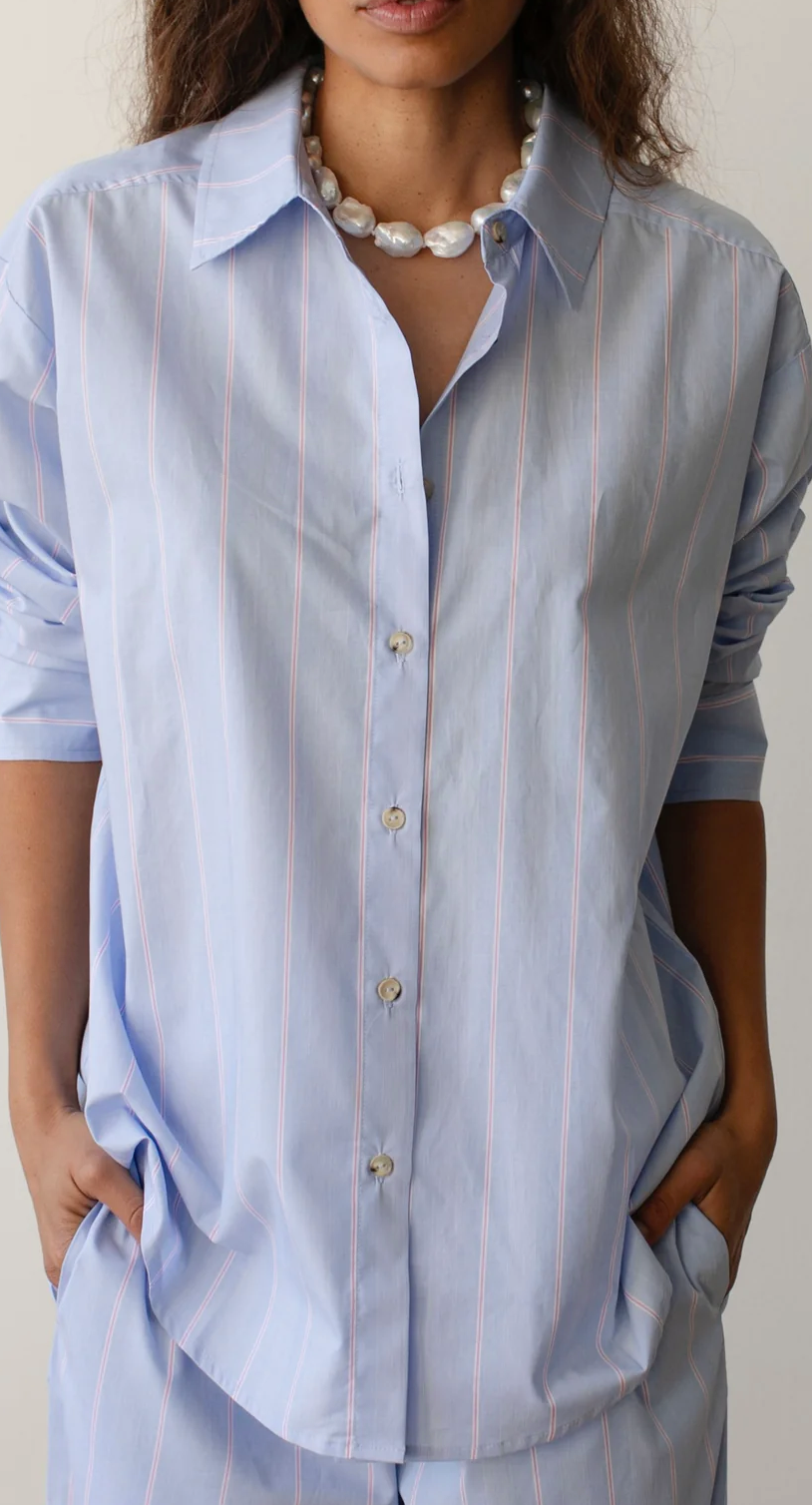 A woman wearing a light blue striped Donni Stripe Pop Shirt with hands on hips. She has a chunky white necklace and the shirt features vertical stripes and wooden buttons.