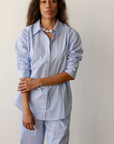 A woman in a light blue Donni Stripe Pop Shirt and matching trousers stands against a plain wall, her arms crossed lightly. She appears serene and confident.
