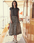 A woman with wavy hair stands on a wooden boat deck. She wears a shadow stripe textured cotton T-shirt, The Viola Skirt by The Great Inc. with a wide smocked waistband, and white sneakers. The boat has a polished wooden cabin door and a shaded canopy area. The background is bright and sunlit.