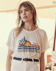 A woman with long, wavy hair stands indoors wearing a white vintage tee featuring a sunset, palm trees, and stripes. She pairs it with high-waisted white pants and a dark belt. With a contemplative expression, she gazes slightly off-camera, showcasing The Boxy Crew by The Great Inc.