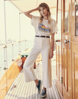 A woman stands on the deck of a boat wearing The Boxy Crew by The Great Inc. with a colorful graphic, belted high-waisted white pants, and black and white sneakers. She is holding her hair with one hand and gazing into the distance. The boat's wooden interior and railing are visible, showcasing her vintage tee style.