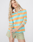 A person with long blonde hair is wearing a collared cotton-blend shirt with orange and teal wide horizontal stripes. The shirt has long sleeves and is paired with matching terry shorts featuring an elastic waist and vertical stripes in the same colors, known as The Terry Venus Short by Kule. The background is plain and light-colored.