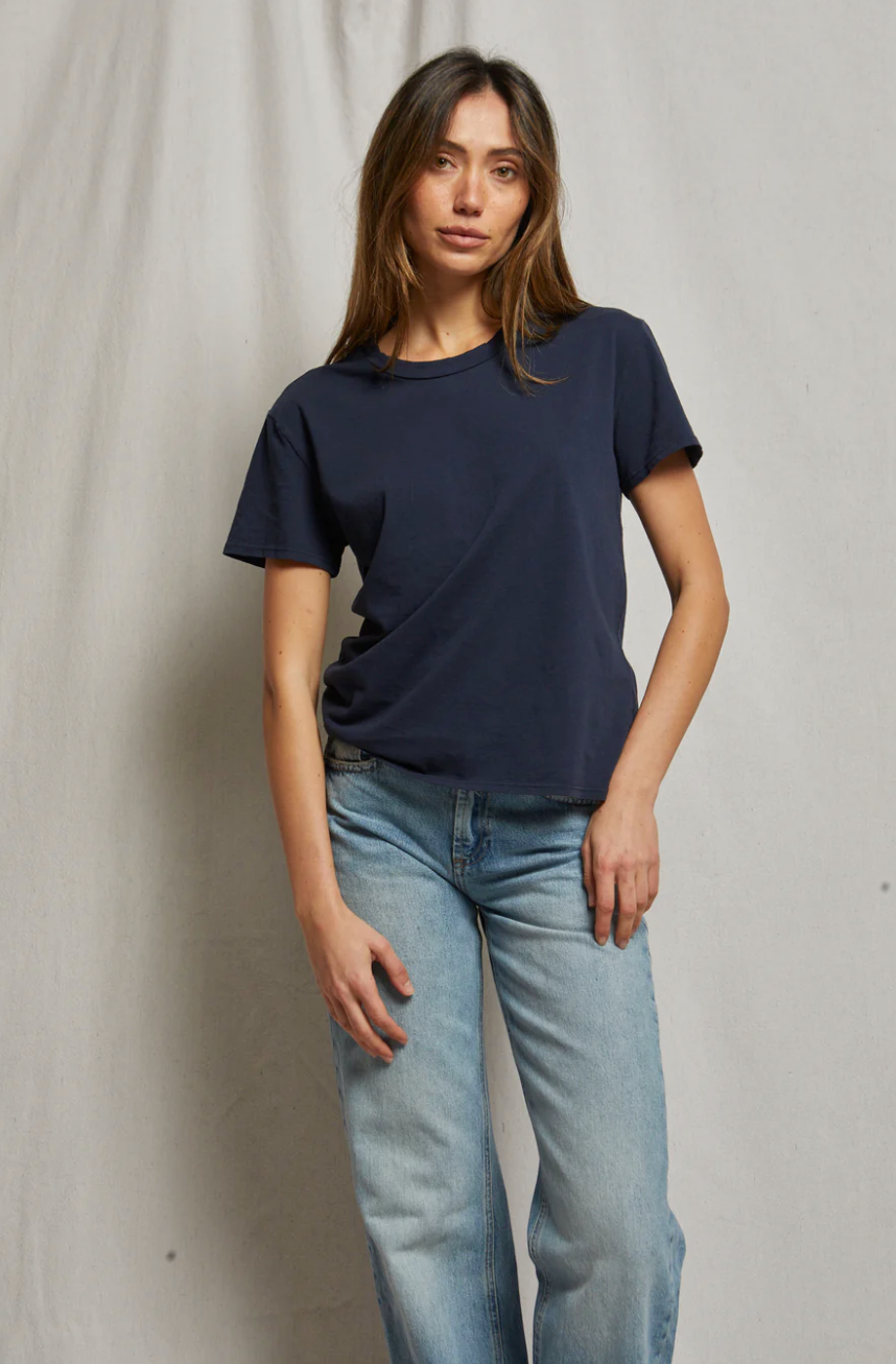A person with long brown hair stands in front of a plain backdrop, exuding a relaxed Arizona style. They are wearing a simple, dark blue Harley SS Boxy Crew from Perfectwhitetee and light blue jeans. Their right hand is resting on the top of their left thigh, and they are looking straight ahead with a neutral expression.