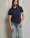 A person with long brown hair stands in front of a plain backdrop, exuding a relaxed Arizona style. They are wearing a simple, dark blue Harley SS Boxy Crew from Perfectwhitetee and light blue jeans. Their right hand is resting on the top of their left thigh, and they are looking straight ahead with a neutral expression.