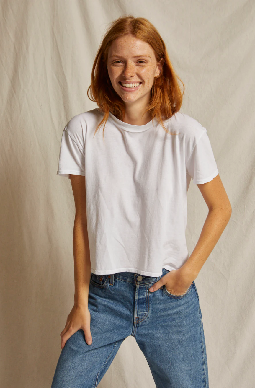 A person with long, red hair smiles while wearing a Perfectwhitetee Harley SS Boxy Crew and high-waisted blue jeans. They have one hand in their pocket and stand against a simple, light-colored background, exuding a relaxed bungalow style.
