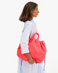 A woman with shoulder-length dark hair is standing sideways, showcasing her style with a bright red Sac Tout from Clare Vivier. She is wearing a light blue long-sleeved dress. The background is white, emphasizing the simplicity and elegance of her look.