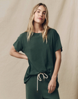 A person sports a loose-fitting, dark green tee reminiscent of a vintage style with matching drawstring pants. They have long, wavy blonde hair and stand against a beige fabric backdrop with one hand on their hip, gazing directly at the camera. The tee they are wearing is The Boxy Crew by The Great Inc.
