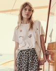 A young woman with long, wavy hair stands leaning against a wooden door on a boat. She is wearing The Boxy Crew by The Great Inc. and leopard print pants that give off vintage tees vibes. The background shows a bright, sunny day on the water.