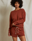 A person with curly hair stands in front of a beige background, wearing a long-sleeved, rust-colored sweatshirt and matching Perfectwhitetee Layla Terry Shorts with an elastic waistband. They are smiling slightly and looking at the camera, with one hand resting on their hip and the other on their wrist.
