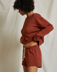 A person stands against an off-white backdrop, adjusting their maroon long-sleeve shirt. They are wearing Perfectwhitetee's Layla Terry Shorts made from French terry cotton, featuring a white drawstring and elastic waistband. With curly hair and a slightly downward gaze, they maintain a relaxed posture.
