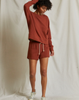 A person with curly hair stands in front of a light, textured backdrop, wearing a matching rust-colored sweatshirt and the Layla Terry Short by Perfectwhitetee, which features an elastic waistband. They have one hand in their hair and look down, smiling. They also wear light-colored sneakers.