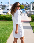 A woman with curly hair, wearing sunglasses and a white Southern Dress from Frank & Eileen, is standing on a pathway by the water in Southern Ireland. She is looking back over her shoulder and smiling. The background includes boats, greenery, and houses.