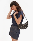 A woman with long dark hair smiles as she looks down. She is wearing a navy short-sleeve button-up shirt with frayed sleeves and a denim skirt. Over her shoulder, she carries the Lune, a black crescent-shaped handbag crafted from chrome tanned leather and adorned with silver daisy studs by Clare Vivier.