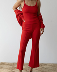 A person is seen wearing a bright red sleeveless top with matching Rib Kick Flare trousers from Donni and a red sweater draped over their shoulders. They have long, curly hair and are accessorized with a pearl necklace. They are standing on a woven carpet with a plain white background.