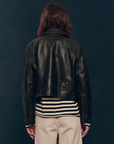 A person with long hair is photographed from behind, wearing The Perfect Leather Jacket by The Great Inc., over a black and white striped shirt and beige pants, standing against a dark background.