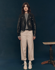A person with shoulder-length brown hair stands against a dark background, sporting The Perfect Leather Jacket from The Great Inc., featuring antique silver snaps. They wear a black and white striped shirt, beige cuffed pants, and white sneakers. Beside them is a wooden stool. The person is looking directly at the camera.