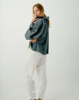 A woman stands with her back slightly turned, wearing the Beautiful Mess Poet Blouse from Aquarius Cocktail, a loose-fitting blue cotton chambray top with a high collar. She pairs it with white, flowy pants. Her long blonde hair is tied in a low ponytail, and she is barefoot. With her left hand touching her chin and a thoughtful expression on her face, she exudes elegance and contemplation.