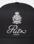 A close-up image of the Ritz Cotton Trucker, a black cotton baseball cap by FRAME, showcases an intricate white embroidered crest with the words "Ritz Paris," adding elegance and a classic touch.