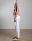 In a minimalist room with a light-colored wall and concrete floor, someone stands sideways wearing Pharaoh's Andy Pant in white, featuring side stripes and an elastic waist, paired with a beige T-shirt and beige sandals.