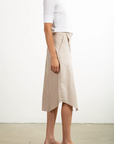 A person is standing in profile against a plain background, wearing a white short-sleeve top and the Pharaoh Nico Skirt with beige and white stripes, made in Los Angeles. They complete their look with beige slide sandals.