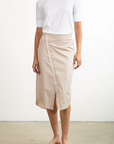 A person stands against a plain background wearing a 100% cotton white T-shirt with Pharaoh's beige striped Nico Skirt featuring an asymmetric placket, completed by chic beige slide sandals.