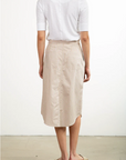 A person is seen from behind wearing a white shirt and the Pharaoh Nico Skirt—a knee-length beige asymmetric placket skirt with vertical stripes and two back pockets. Paired with beige flat sandals, this chic outfit stands out against an off-white wall background. Proudly made in Los Angeles.