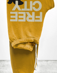 The FREECITY Large Sweatpant by Free City (sparrow, LLC) in yellow is a unisex design crafted in the USA from lightweight French terry. It features an elastic waistband with a white drawstring, "FREE CITY" text, and a bird silhouette. Displayed elegantly on a light gray surface.