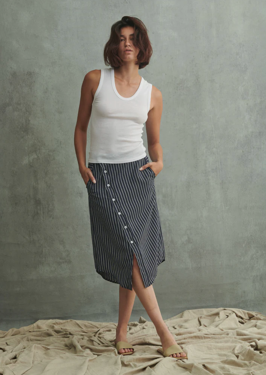 In Los Angeles, a person with short hair stands against a textured concrete background, wearing a white sleeveless top and Pharaoh's Nico Skirt featuring an asymmetric placket adorned with buttons. Their hands rest in the front slash pockets of the skirt, and they are barefoot on the draped fabric floor.