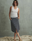 In Los Angeles, a person with short hair stands against a textured concrete background, wearing a white sleeveless top and Pharaoh's Nico Skirt featuring an asymmetric placket adorned with buttons. Their hands rest in the front slash pockets of the skirt, and they are barefoot on the draped fabric floor.