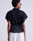 A person with short hair wearing sunglasses and an Agni Floaty Top by Apiece Apart, made from organic cotton, is paired with white pants. They face away from the camera against a plain backdrop, forming an intriguing silhouette.